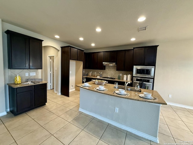 kitchen with sink, light stone counters, light tile patterned floors, appliances with stainless steel finishes, and an island with sink