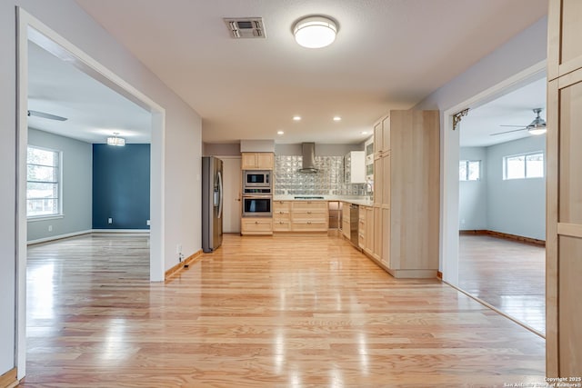 kitchen featuring ceiling fan, appliances with stainless steel finishes, tasteful backsplash, wall chimney exhaust hood, and light wood-type flooring