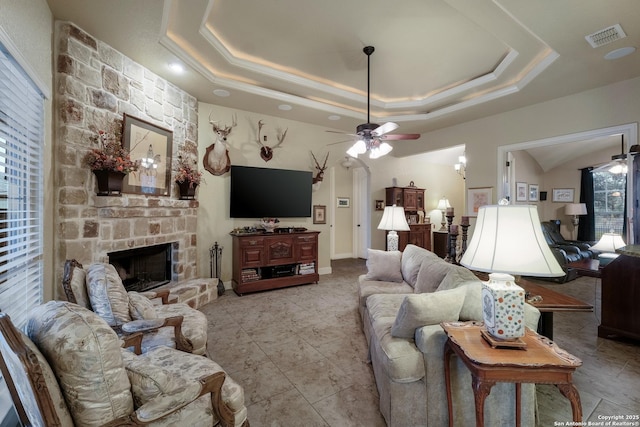 living room featuring crown molding, a tray ceiling, a stone fireplace, and ceiling fan