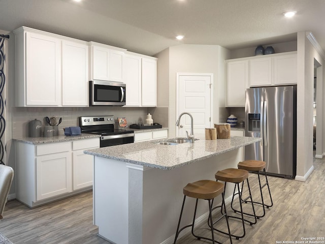 kitchen with light stone counters, an island with sink, a sink, appliances with stainless steel finishes, and light wood-type flooring