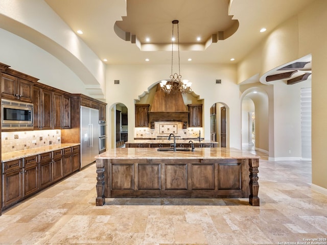 kitchen featuring sink, built in appliances, a large island with sink, a high ceiling, and backsplash