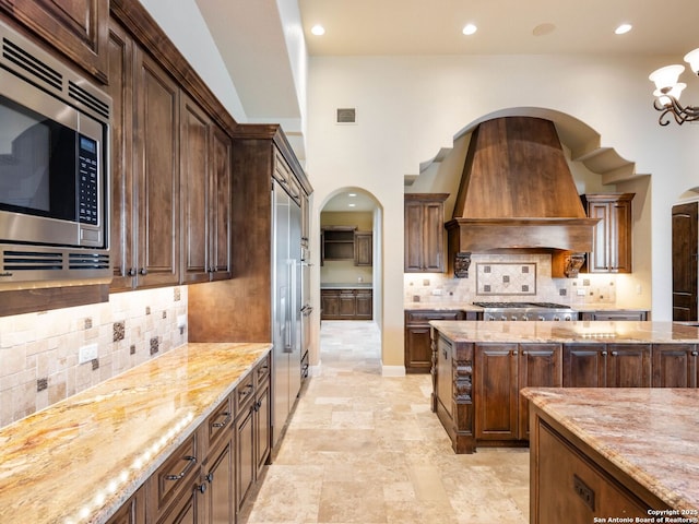 kitchen featuring premium range hood, backsplash, dark brown cabinets, stainless steel microwave, and light stone countertops