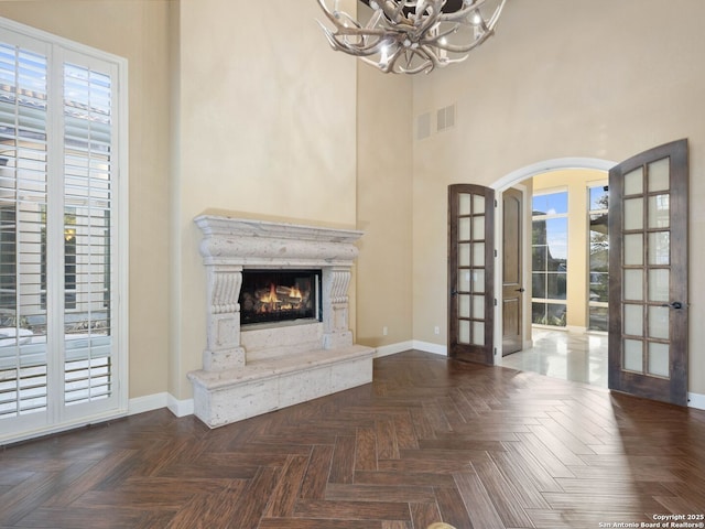 unfurnished living room with a towering ceiling, a chandelier, dark parquet flooring, a high end fireplace, and french doors