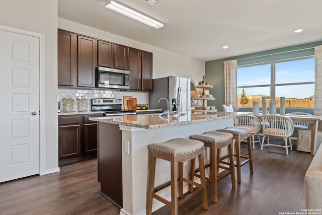 kitchen featuring appliances with stainless steel finishes, an island with sink, a kitchen breakfast bar, dark hardwood / wood-style flooring, and light stone countertops