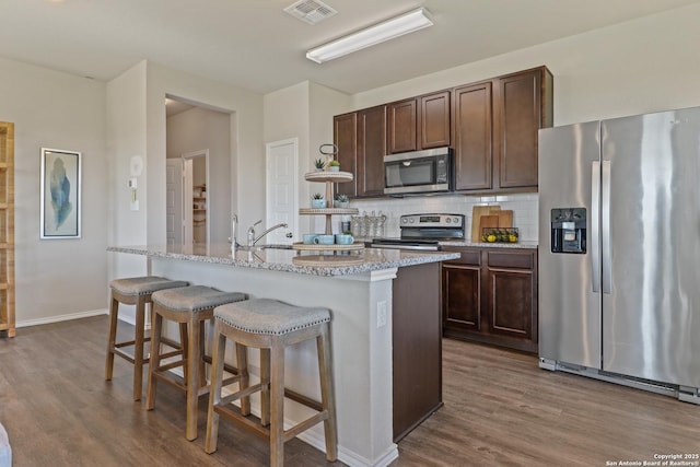 kitchen with dark brown cabinetry, light stone counters, an island with sink, stainless steel appliances, and backsplash