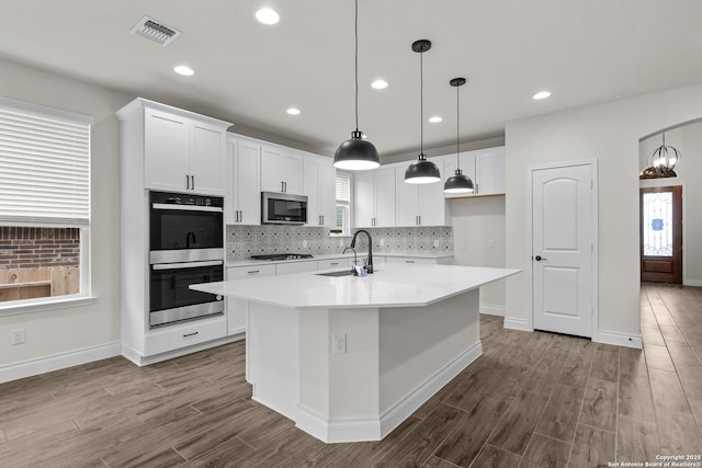 kitchen with sink, a kitchen island with sink, stainless steel appliances, white cabinets, and decorative light fixtures