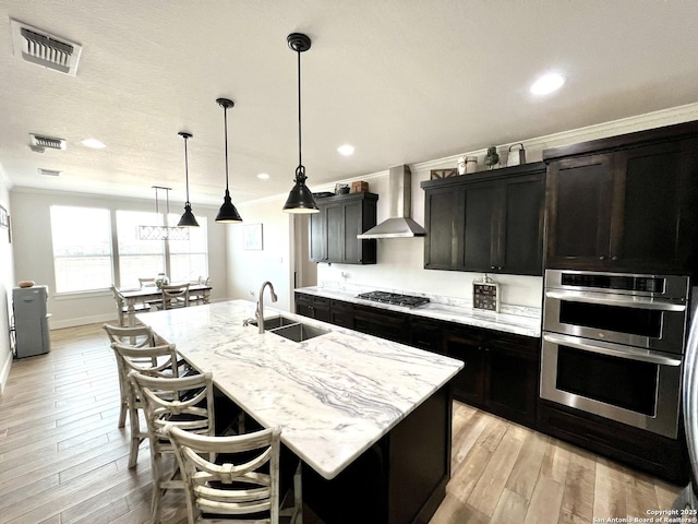 kitchen featuring appliances with stainless steel finishes, an island with sink, sink, ornamental molding, and wall chimney exhaust hood