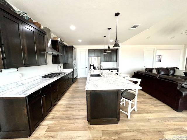 kitchen with decorative light fixtures, sink, wall chimney range hood, light hardwood / wood-style floors, and a center island with sink