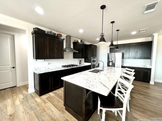 kitchen featuring appliances with stainless steel finishes, sink, hanging light fixtures, a kitchen island with sink, and wall chimney range hood