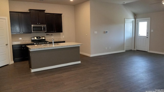 kitchen featuring sink, dark wood-type flooring, stainless steel appliances, light stone countertops, and a center island with sink