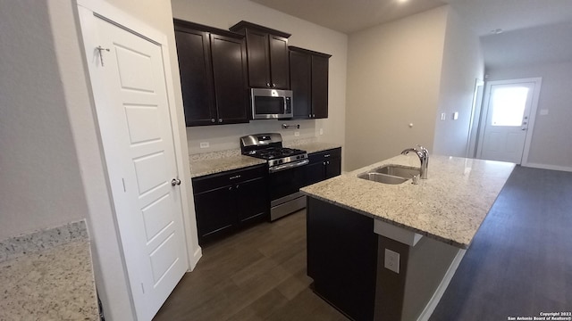 kitchen with appliances with stainless steel finishes, sink, a kitchen island with sink, light stone countertops, and dark wood-type flooring