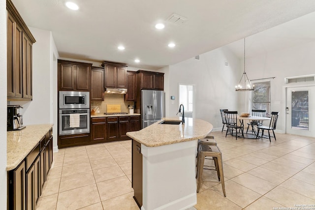 kitchen featuring sink, decorative light fixtures, appliances with stainless steel finishes, light stone countertops, and a kitchen island with sink