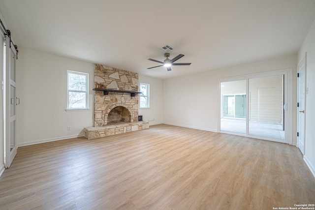 unfurnished living room with a stone fireplace, a barn door, ceiling fan, and light wood-type flooring