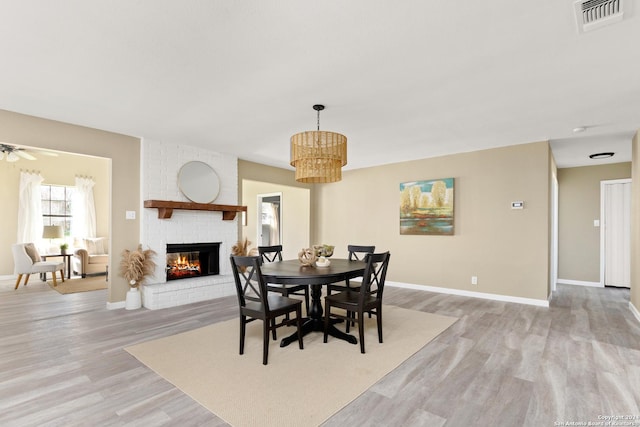 dining area featuring ceiling fan with notable chandelier, a brick fireplace, and light wood-type flooring