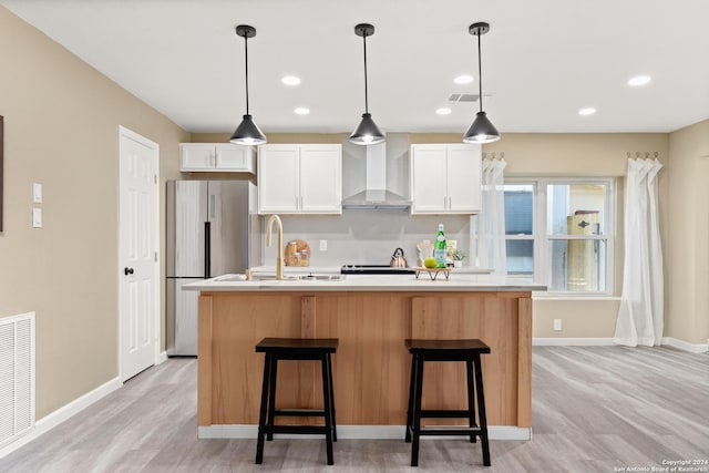 kitchen featuring a kitchen island with sink, wall chimney range hood, stainless steel refrigerator, and white cabinets