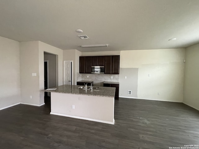 kitchen with tasteful backsplash, light stone counters, dark brown cabinets, a center island with sink, and dark hardwood / wood-style flooring