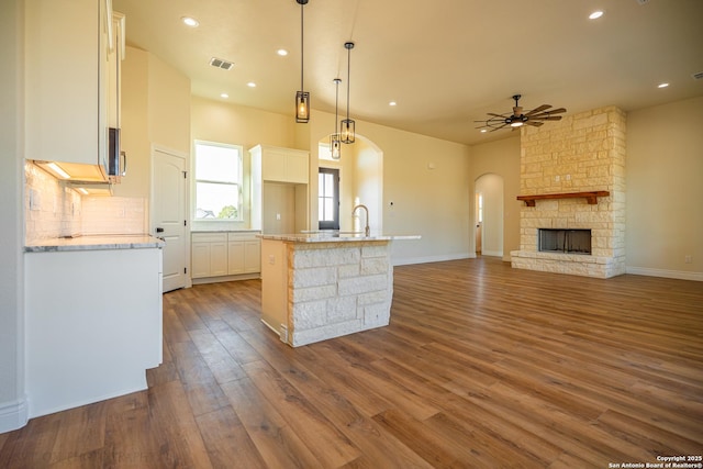 kitchen with a stone fireplace, pendant lighting, white cabinets, a kitchen island with sink, and ceiling fan