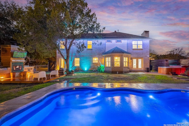 pool at dusk with a hot tub, a patio, exterior fireplace, and a lawn