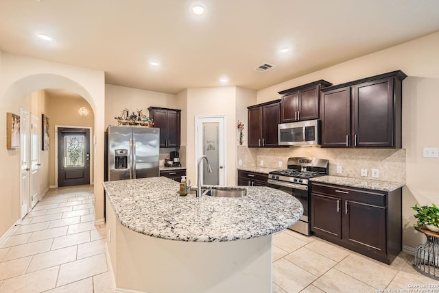 kitchen with sink, light tile patterned floors, appliances with stainless steel finishes, light stone counters, and an island with sink