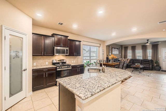 kitchen with sink, appliances with stainless steel finishes, a kitchen island with sink, dark brown cabinets, and light stone counters