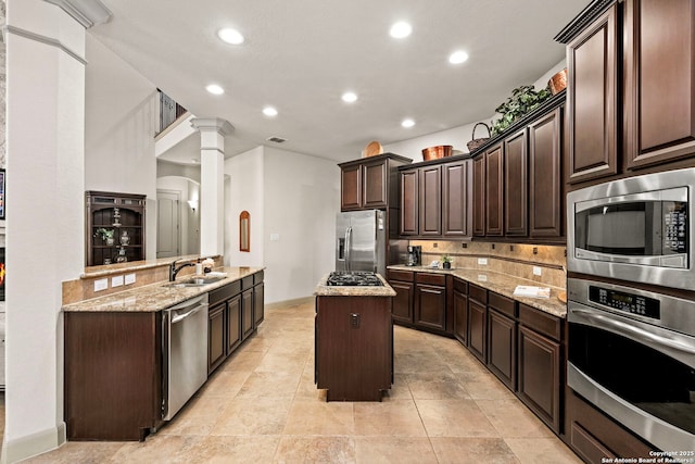 kitchen featuring decorative columns, appliances with stainless steel finishes, dark brown cabinetry, a kitchen island, and a sink