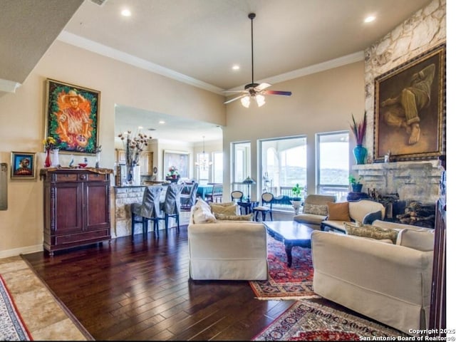 living room with dark wood-type flooring, ceiling fan with notable chandelier, a towering ceiling, ornamental molding, and a stone fireplace