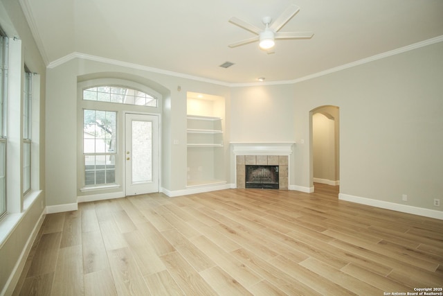 unfurnished living room featuring built in shelves, light wood-type flooring, ornamental molding, a tile fireplace, and ceiling fan