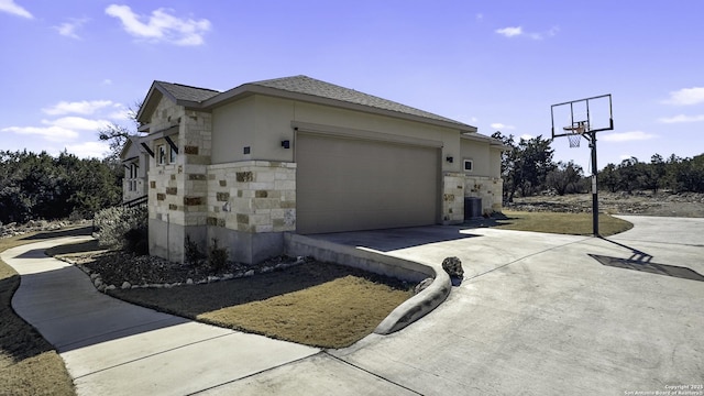 view of side of home with driveway, stone siding, roof with shingles, an attached garage, and cooling unit