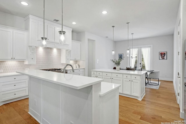 kitchen with pendant lighting, a kitchen island with sink, backsplash, light hardwood / wood-style floors, and white cabinets