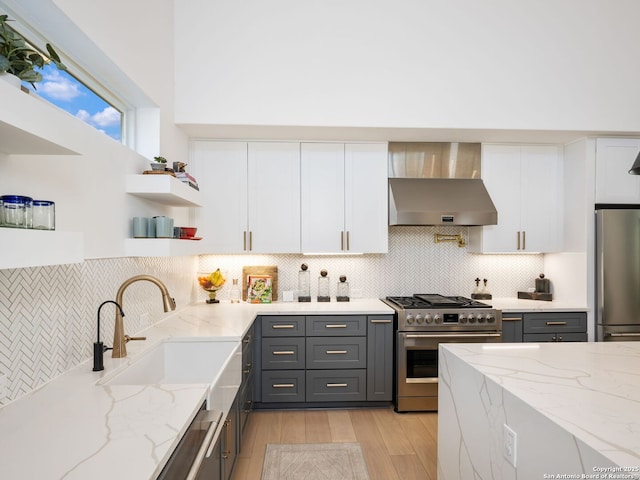 kitchen with wall chimney range hood, gray cabinetry, stainless steel appliances, light stone countertops, and white cabinets