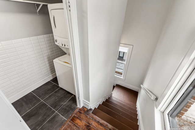 laundry room with dark tile patterned floors, stacked washer / drying machine, and tile walls