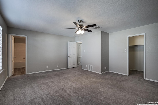 unfurnished bedroom featuring ensuite bathroom, a walk in closet, a textured ceiling, dark carpet, and ceiling fan