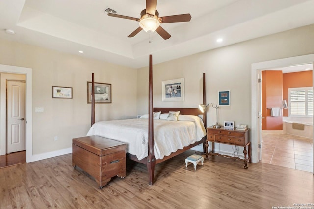 bedroom with ceiling fan, connected bathroom, a tray ceiling, and light hardwood / wood-style flooring