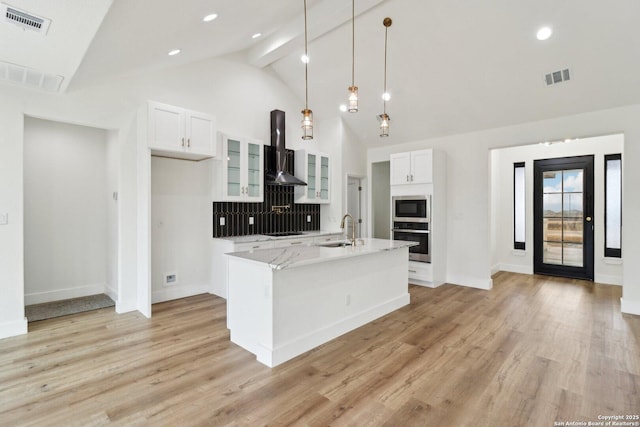 kitchen with an island with sink, white cabinets, stainless steel oven, light stone countertops, and wall chimney exhaust hood