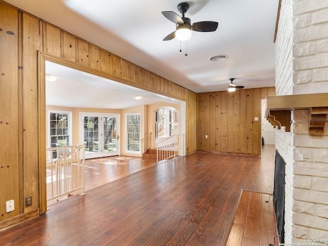 unfurnished living room featuring dark hardwood / wood-style flooring, a stone fireplace, ceiling fan, and wood walls