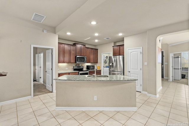 kitchen featuring light tile patterned flooring, backsplash, a kitchen island with sink, stainless steel appliances, and light stone countertops