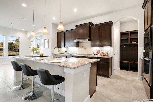 kitchen featuring a center island with sink, appliances with stainless steel finishes, pendant lighting, light stone countertops, and decorative backsplash