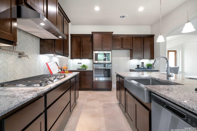 kitchen featuring appliances with stainless steel finishes, decorative light fixtures, sink, light stone counters, and wall chimney range hood