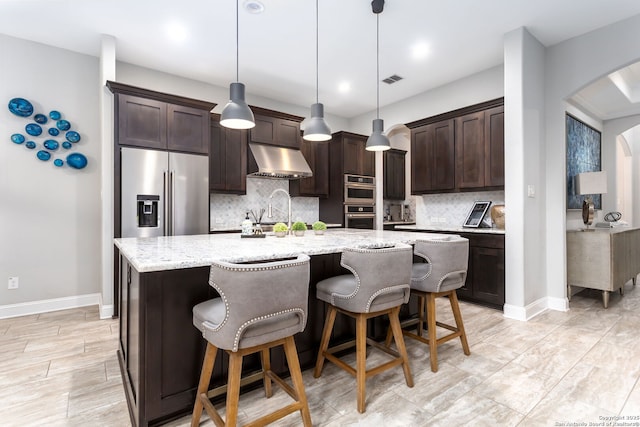 kitchen with decorative light fixtures, stainless steel appliances, light stone countertops, dark brown cabinets, and a center island with sink