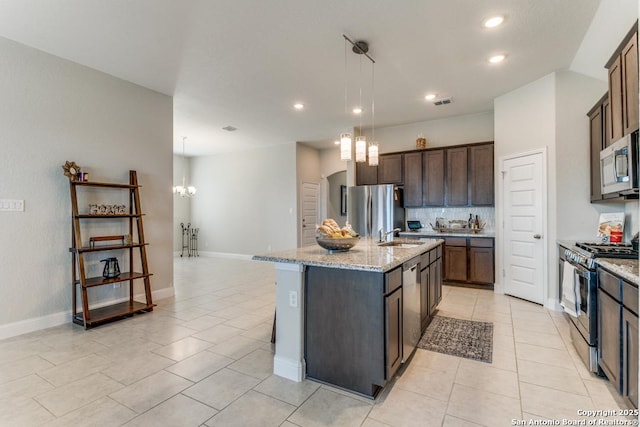 kitchen with pendant lighting, appliances with stainless steel finishes, dark brown cabinets, light stone countertops, and an island with sink