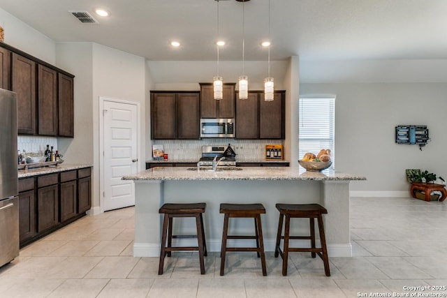 kitchen featuring a kitchen island with sink, hanging light fixtures, light stone countertops, and appliances with stainless steel finishes