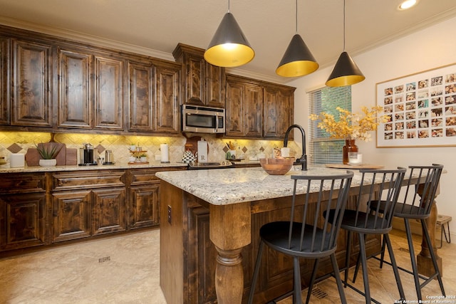 kitchen featuring a kitchen island with sink, light stone counters, tasteful backsplash, dark brown cabinetry, and ornamental molding