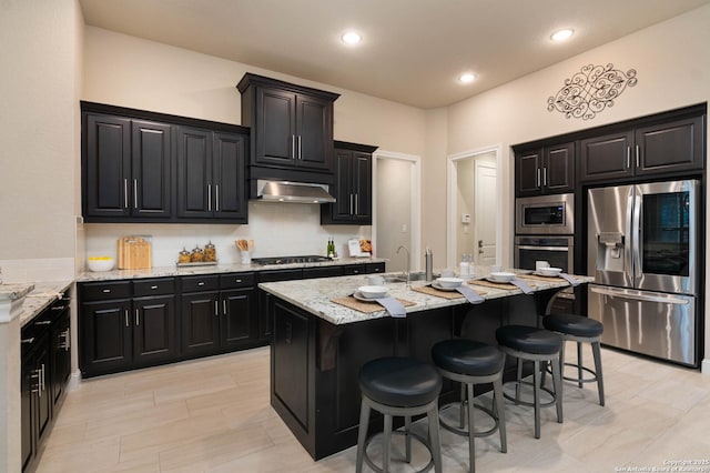 kitchen with a breakfast bar, decorative backsplash, stainless steel appliances, light stone countertops, and a center island with sink