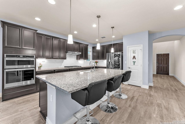 kitchen with light stone counters, hanging light fixtures, a kitchen breakfast bar, stainless steel appliances, and a kitchen island with sink