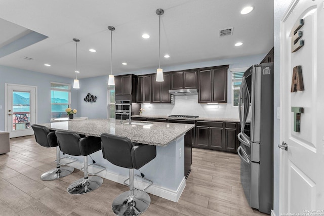 kitchen featuring appliances with stainless steel finishes, pendant lighting, a kitchen bar, light stone counters, and a center island with sink