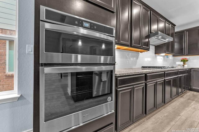 kitchen featuring backsplash, light stone counters, stainless steel appliances, dark brown cabinets, and light hardwood / wood-style flooring