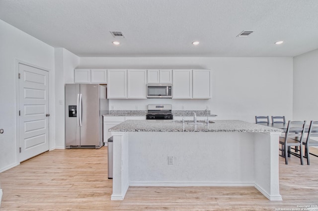 kitchen featuring sink, white cabinets, stainless steel appliances, light stone countertops, and a center island with sink