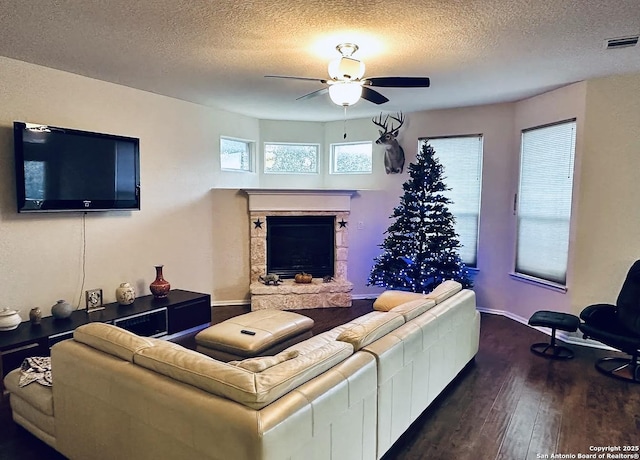 living room with a textured ceiling, dark wood-type flooring, a stone fireplace, and ceiling fan