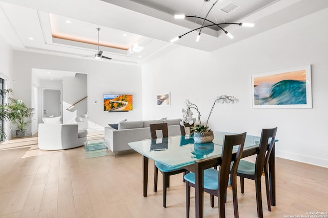 dining space featuring a raised ceiling, a high ceiling, ceiling fan with notable chandelier, and light wood-type flooring