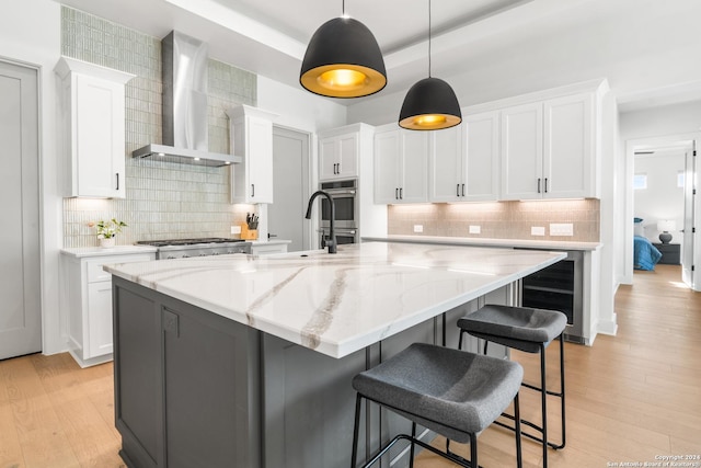 kitchen featuring a large island with sink, wall chimney range hood, white cabinets, and decorative light fixtures